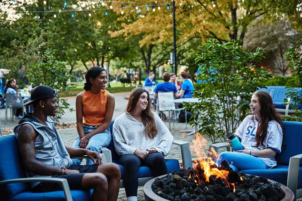 Four students sit around a lit firepit ourside of a SLU residence hall