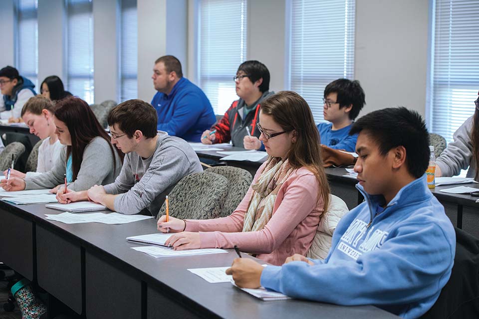 Students holding pens and pencils sit along a long classroom table, writing on pieces of paper.