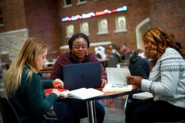 Three female students work at a round table in the atrium of the business school. Two students work in books and one is working on a laptop.