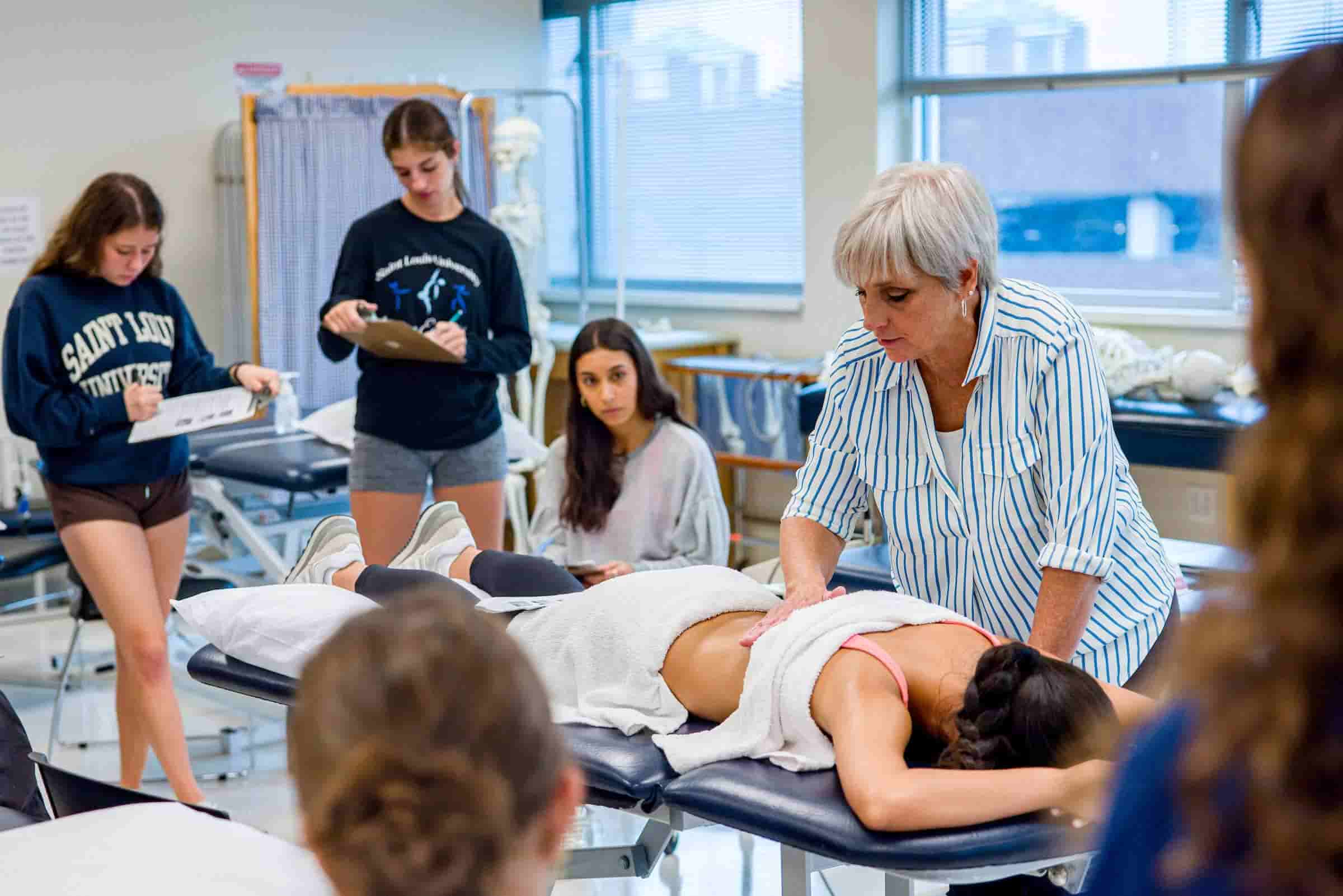 A female professor demonstrates physical therapy techniques on a real model in front of a class of students. The model is lying face down on a massage bed.