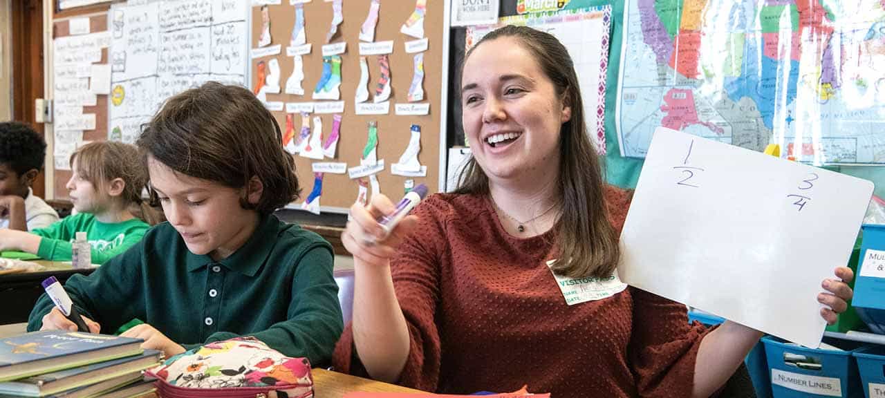 A female SLU student sits on the floor of an elementary school classroom. Young students sit in a circle around her as she holds up a flashcard.