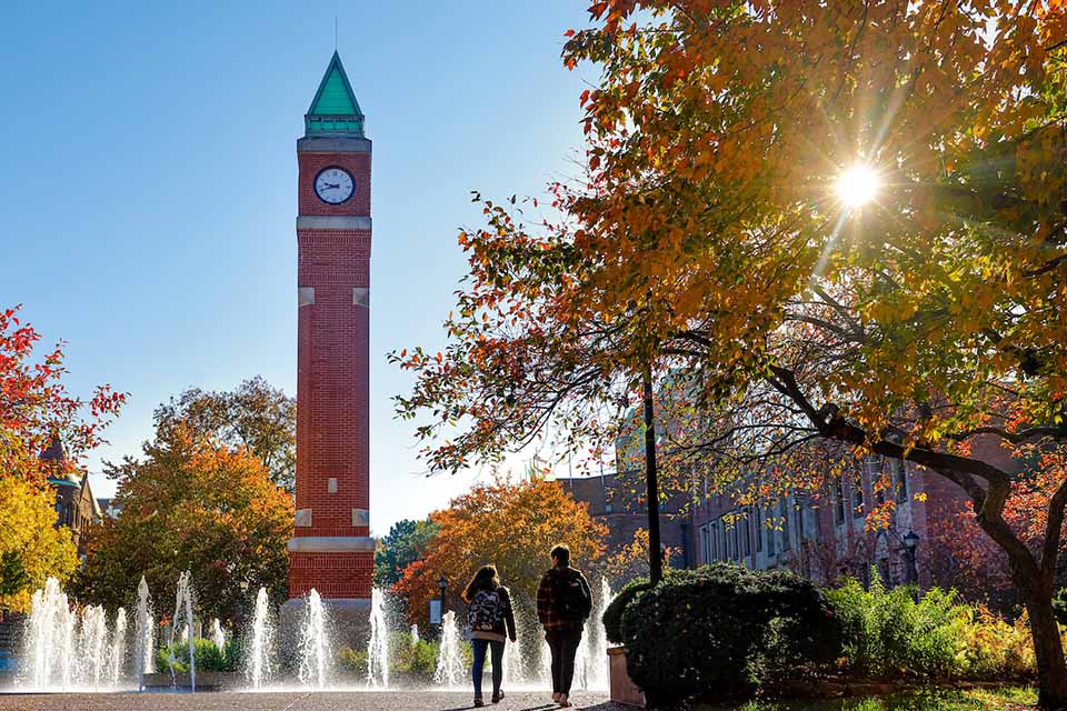 A clocktower on campus with water fountains surrounding it and two students walking toward it.