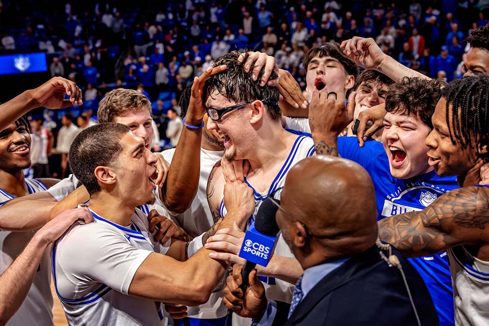 A men's basketball player is surrounded by teammates and someone with a microphone while celebrating on the court.