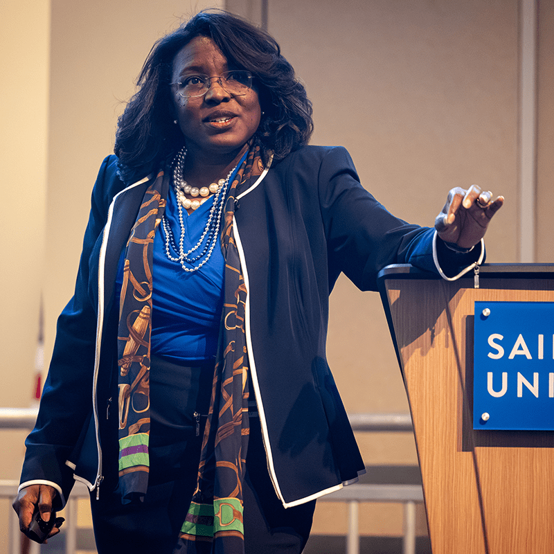 A woman in a professional suit stands next to a lectern during a lecture.