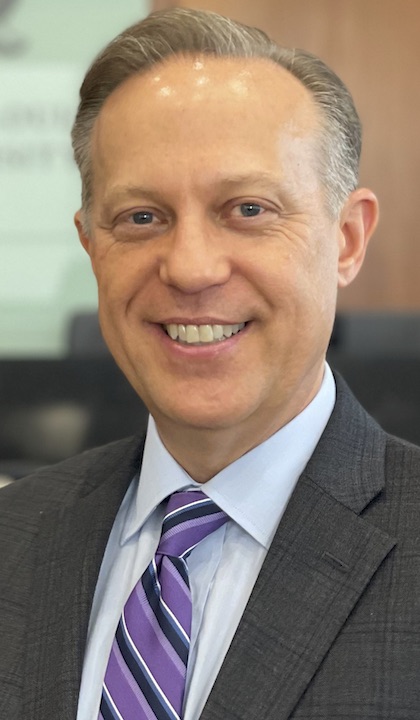 Headshot of Matthew Tuegel, wearing dark suit, blue shirt and purple striped tie.