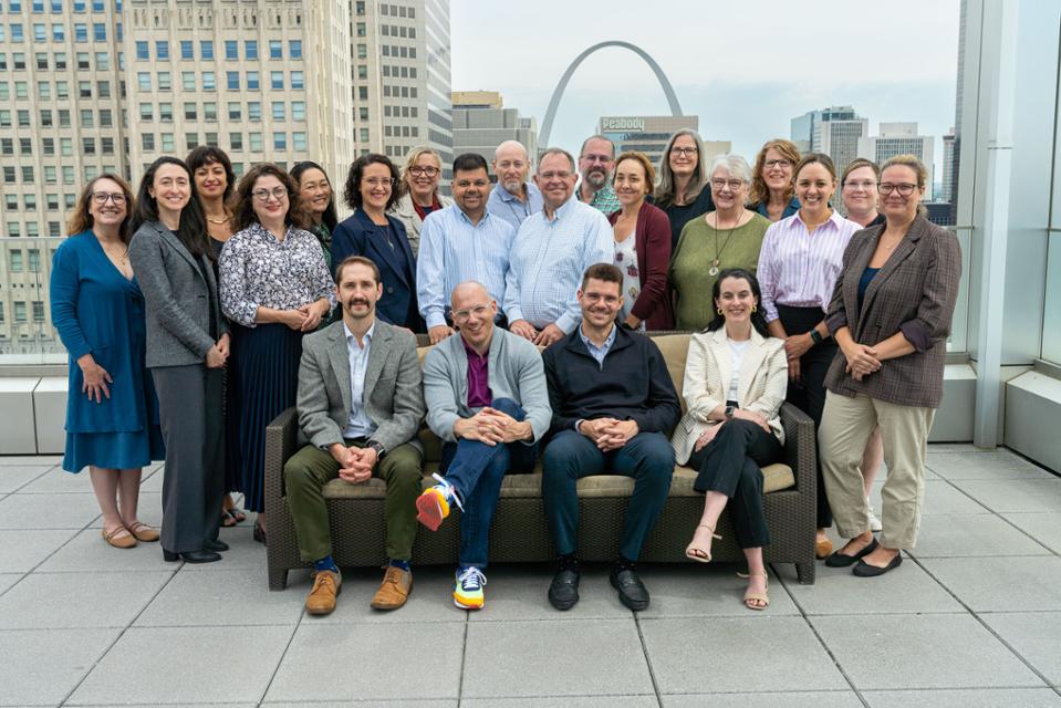 Center Health Law Studies faculty, staff and scholars pose in a group in the roof top pavilion of Scott Hall in St. Louis. A group of people pose for a photo on a rooftop patio with the St. Louis Arch in the background