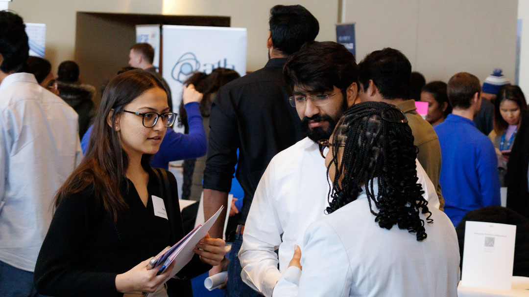 Two students talk to an employer in a room filled with people.
