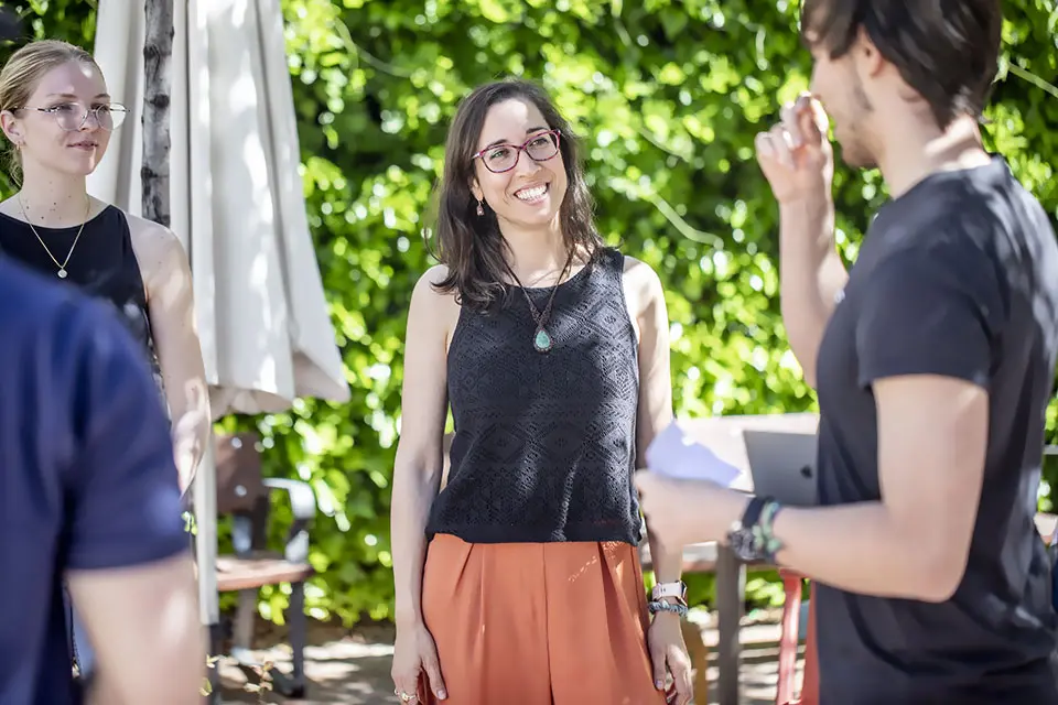 Students converse while standing outside under some trees on a sunny day.