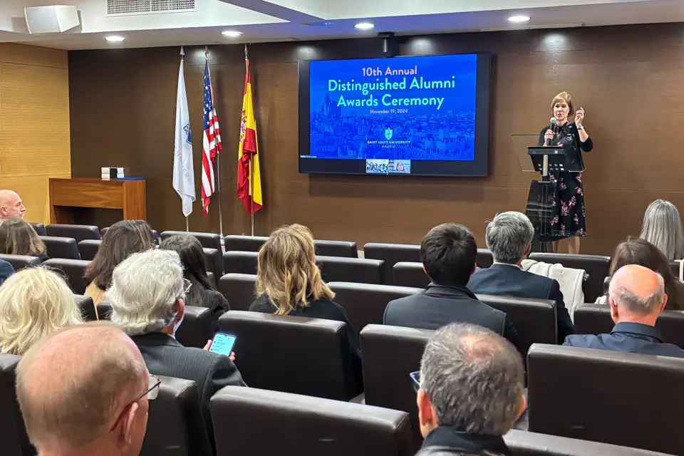 Woman with a microphone standing behind a lectern welcomes the audience in the auditorium with the alumni ceremony poster on screen.