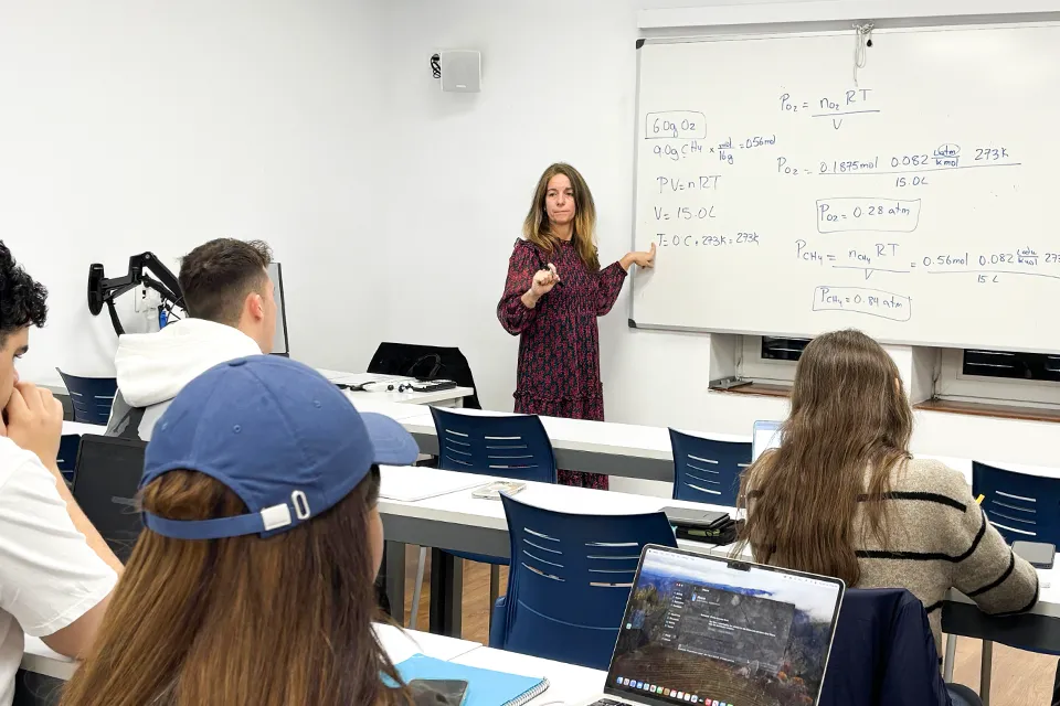 Woman in front of class while pointing to an equation on the whiteboard behind her with a computer monitor next to her while students look toward the front of the room.