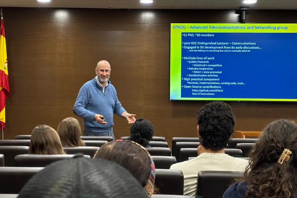 A professor stands at the front of a classroom delivering a presentation on advanced telecommunications and networking to a seated audience of students, with a slide displayed on a screen and a Spanish flag visible to the side.
