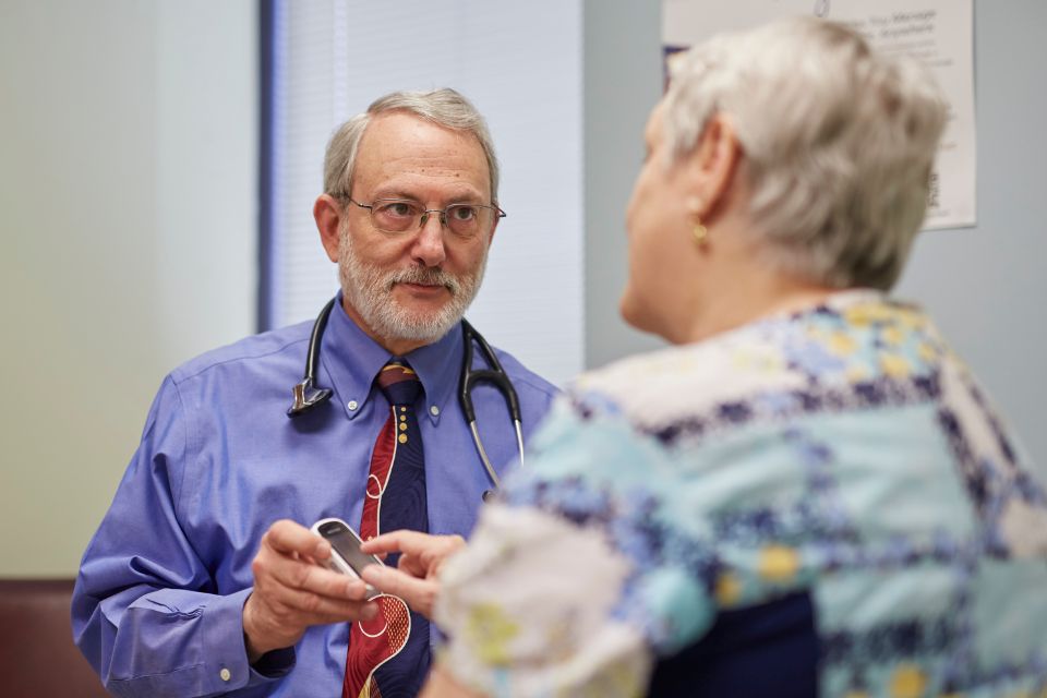 An endocrinologist holding a blood sugar monitoring device speaks with a patient inside a hospital exam room.