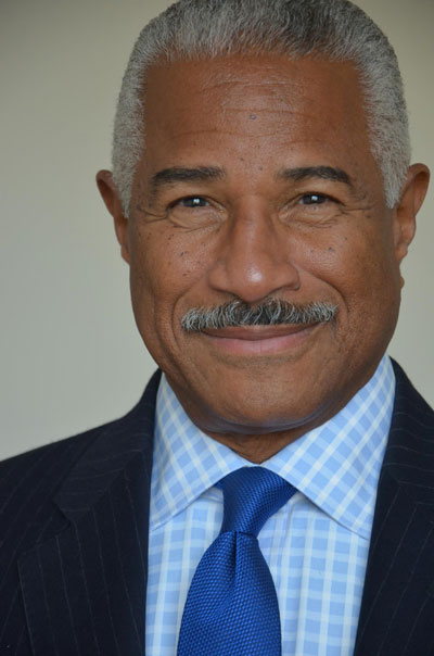 A close-up photo of poet and activist Felipe Luciano, dressed in pale blue-and-white checkered shirt, cobalt blue tie and navy blue pinstripe jacket