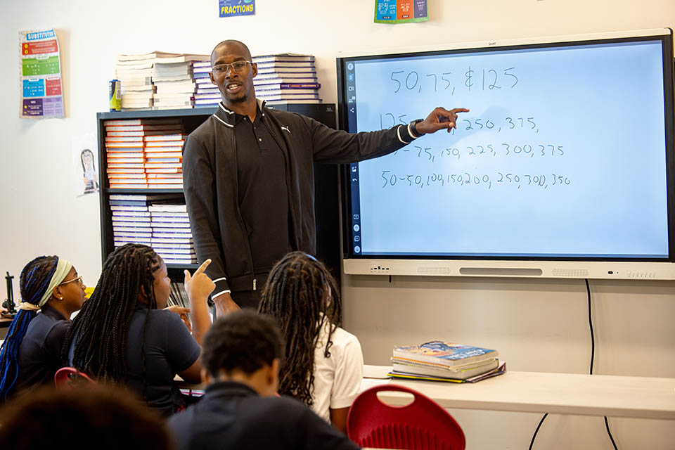 A man instructs a class. He points to a white board with numbers written on it. 