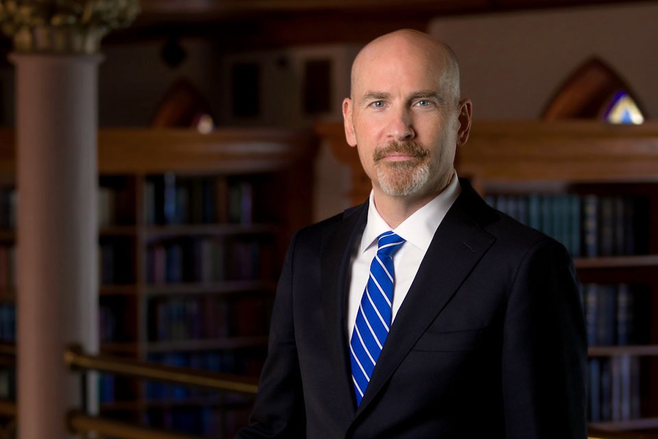 SLU President-Elect Edward J. Feser, wearing a blue suit and tie, stands before a collection of bookshelves, showcasing a variety of books. SLU President-Elect Edward J. Feser, wearing a dark suit and royal blue tie, stands before a collection of bookshelves, showcasing a variety of books.