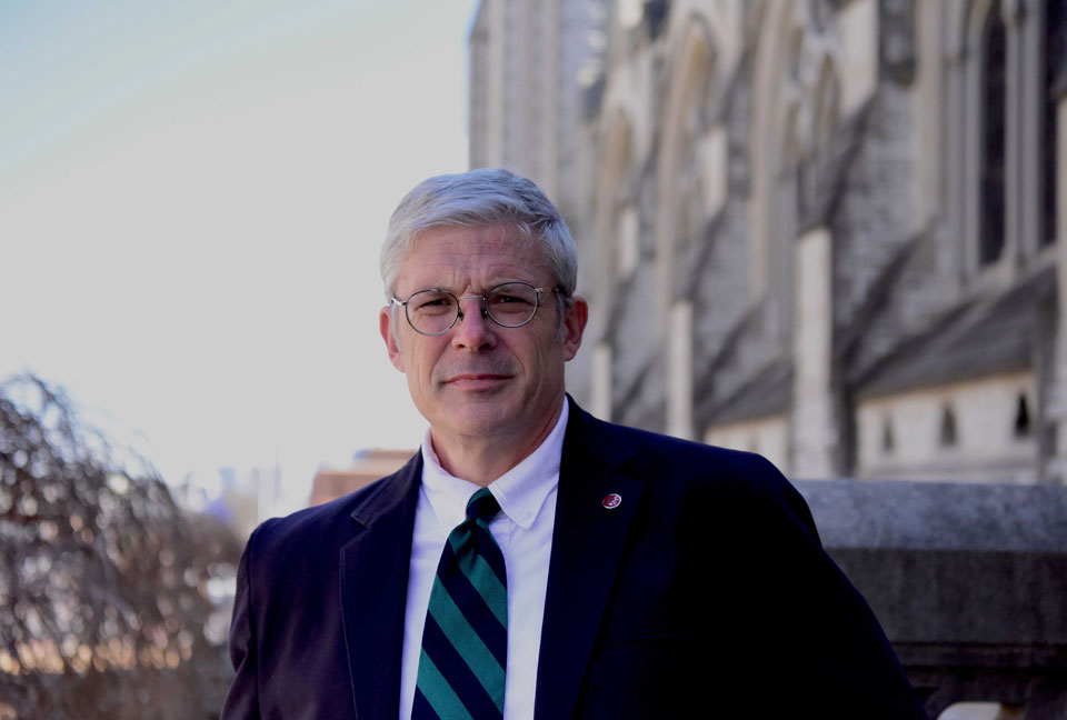 A cropped photo of Dr. Thomas Finan from the waist up. He poses for a photo outdoors during the day. A building is in the background on his left.