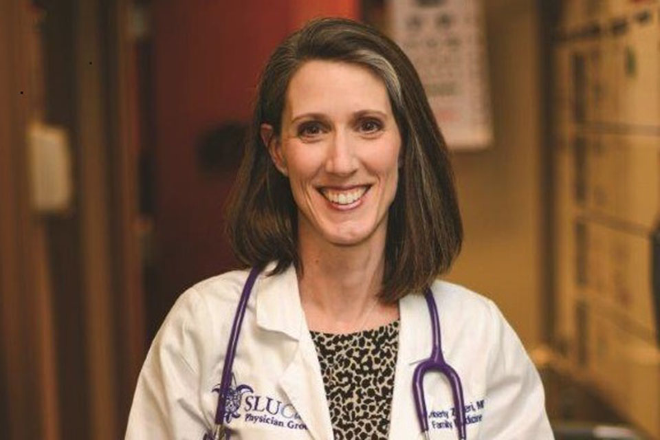 Kimberly Schiel, M.D. Dr. Kimberly Schiel poses for a photo in a dimly lit room in a white coat and a stethoscope around her neck.