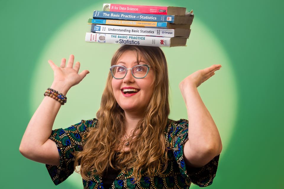 A woman balances five books on her head. Her hands are raised as if to catch the books if they were to fall. She is smiling and wearing glasses. She takes a photo against a green background with light green spotlight.