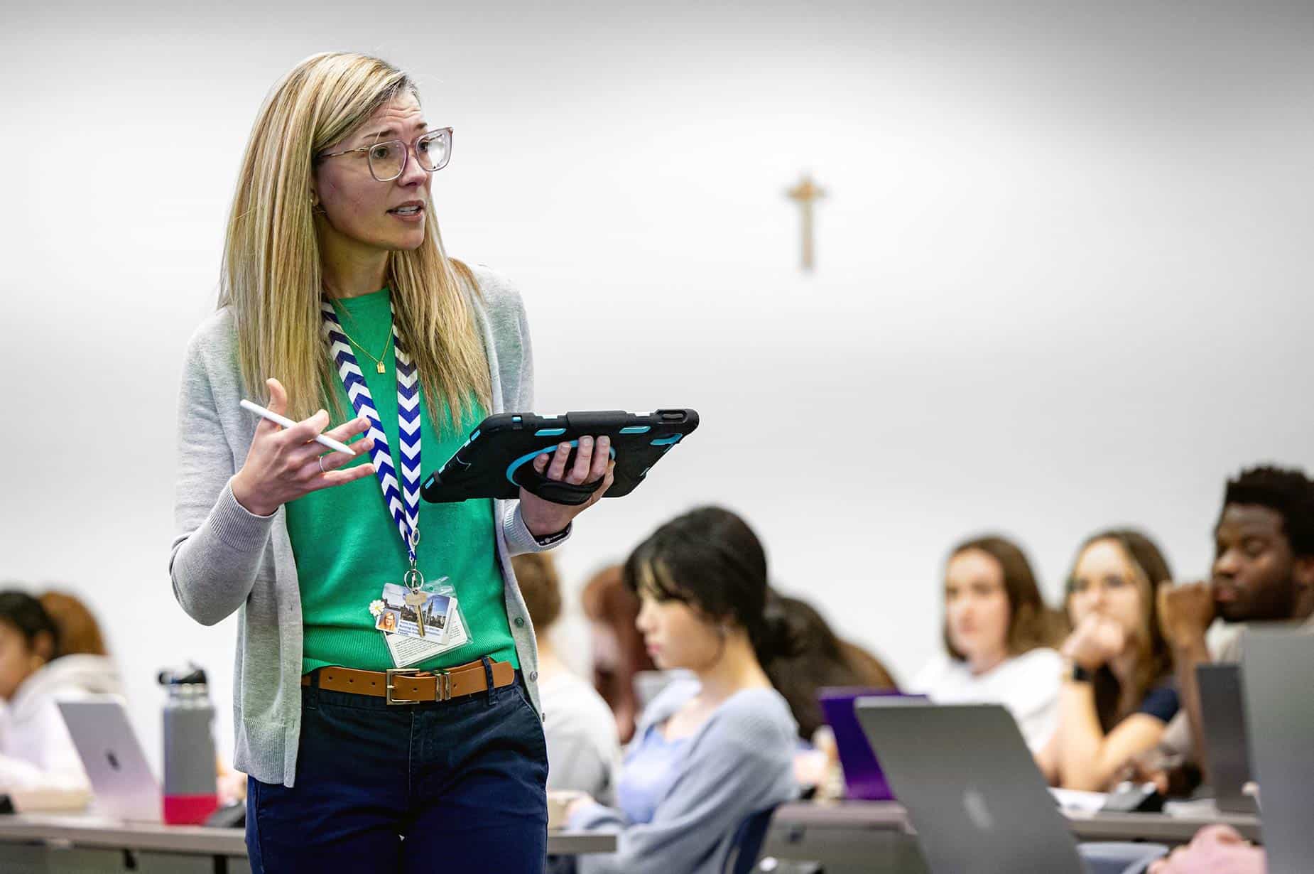 A nursing professor, wearing a green blouse and holding a tablet, speaks to a classroom full of students. Students of different genders and races can be seen in the background working on laptops at classroom desks.