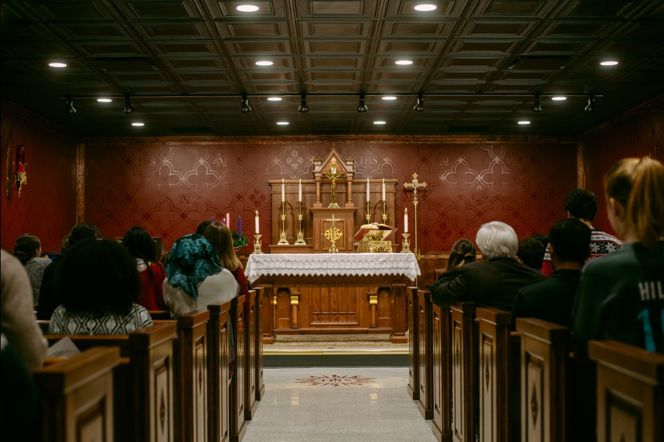 The Catholic Studies Centre Chapel People sit, seen from behind, in the Catholic Studies Centre Chapel, looking at the altar. The altar is made of wood with a table covered in a lace cloth and candles, crosses and other accessories in gold.