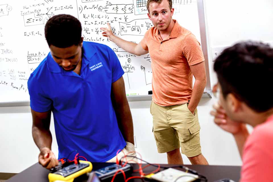 A man gestures toward a white board with formulas written on it