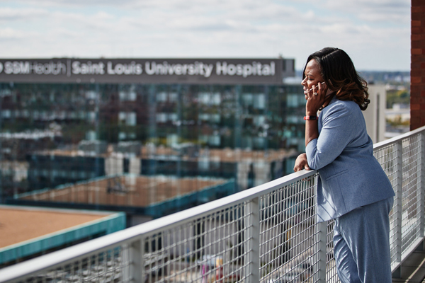 A health administration professional takes a work phone call on the balcony of an office building with the Saint Louis University Hospital in the background on a sunny day in the afternoon.