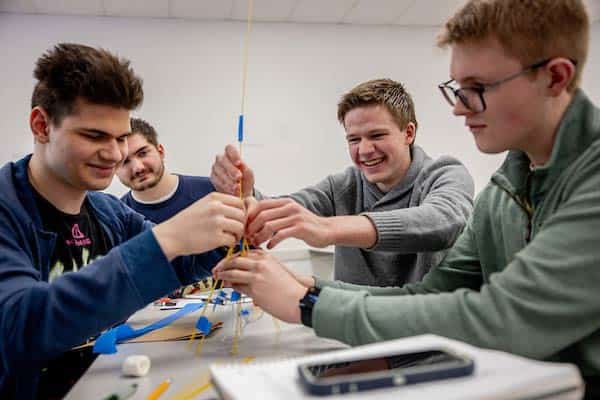 A group of four male students work together to construct a tower out of raw spaghetti and blue painter’s tape in a classroom.