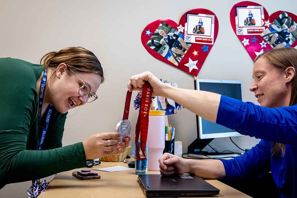 Sarah Adam shows off her silver Paralympic medal to a SLU colleague in her office 