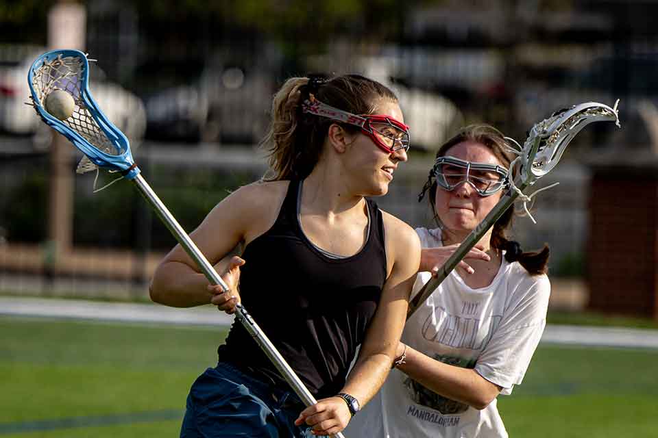 Two female lacrosse players take part in a game on the field on a sunny day.