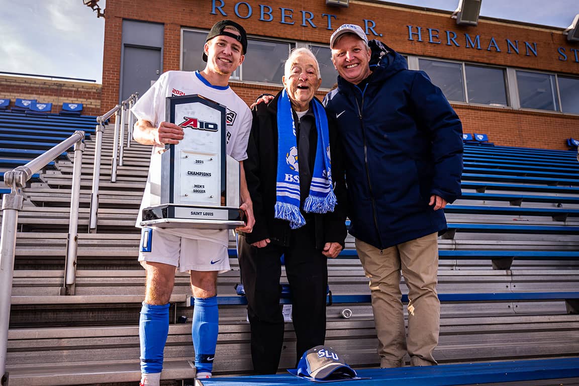 An elderly man, a middle aged man and a young man — the Kleins — pose for a photo with a men's A-10 Championship trophy in the stands of Hermann Stadium.