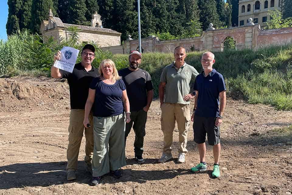 A group of researchers stand near the Italian dig site. One male researcher holds up a sign that reads "Saint Louis University."