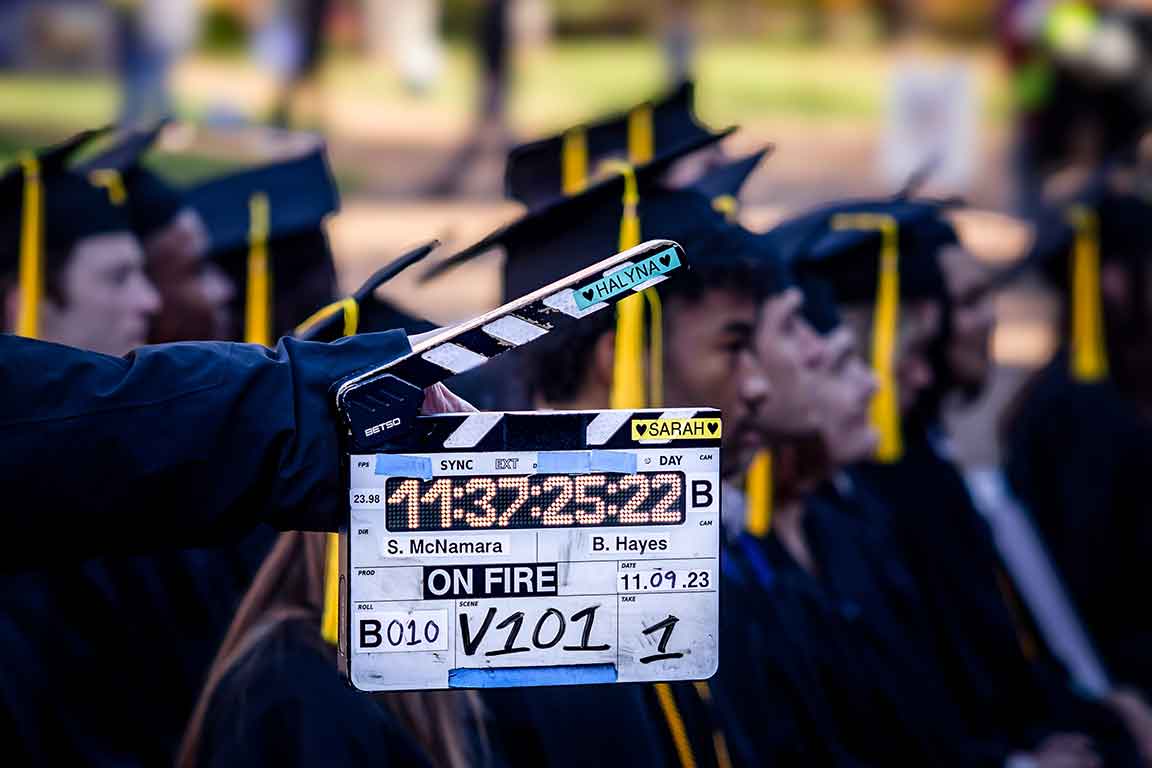 A clapperboard with the words "ON FIRE" is positioned in front of a crowd of actors wearing graduation caps and gowns.